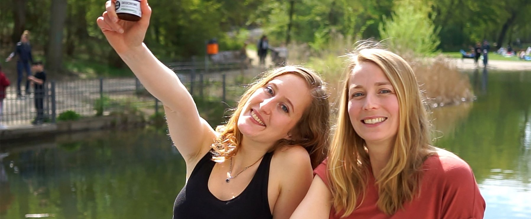 Zwei lächelnde Frauen sitzen an einem Teich. Eine Frau hält ein kleines Gefäß hoch. Im Hintergrund Bäume und Menschen.