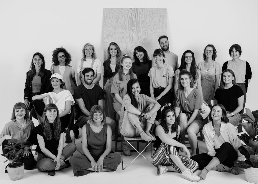 Eine Gruppe Erwachsener in Freizeitkleidung posiert für ein Schwarzweißfoto in einem Studio mit einer Holzkulisse im Hintergrund und zwei Topfpflanzen im Vordergrund.