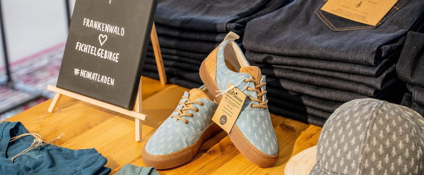 Lässige Sneakers mit dekorativem Aufdruck werden neben gefalteten Jeans, einer Mütze, einem Hemd und einem kleinen Schild mit Text auf einem Holztisch präsentiert.
