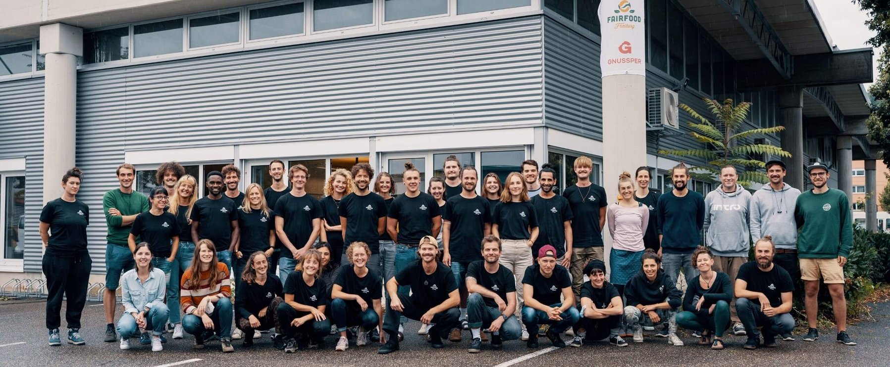 Eine Gruppe von Menschen in legerer Kleidung posiert für ein Foto vor einem Gebäude mit Lamellenfassade und einem Schild mit der Aufschrift „Fairfood“ und „Gnussper“.
