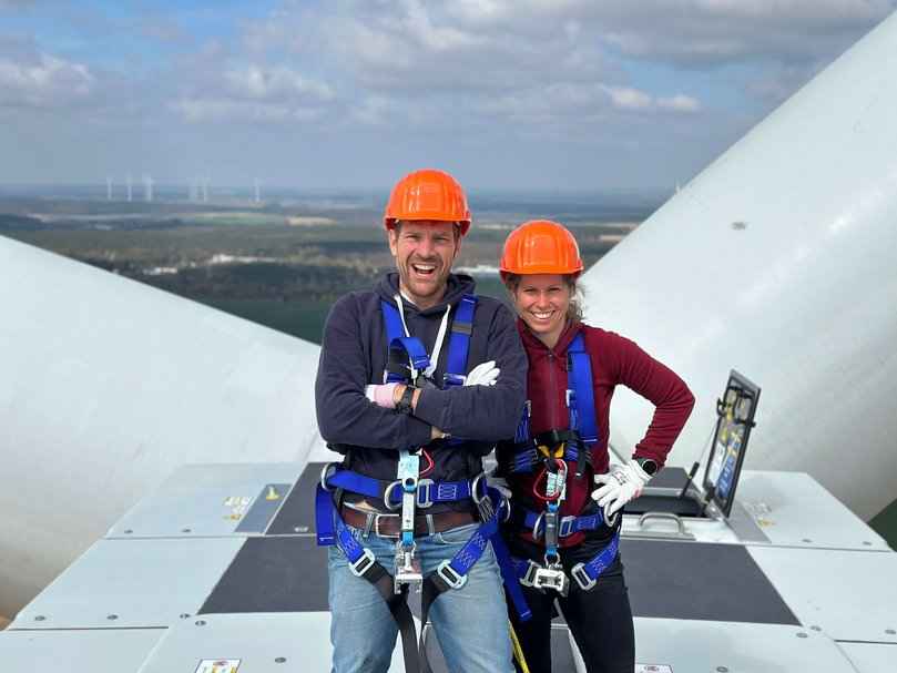 Zwei Personen mit Sicherheitsgurten und orangefarbenen Helmen stehen auf einer Windkraftanlage mit großen Rotorblättern und einer Landschaft im Hintergrund.