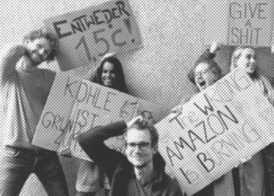Eine Gruppe von vier Personen hält Protestschilder mit verschiedenen Botschaften hoch, darunter eines mit der Aufschrift „Entweder 1,5 °C!“ und ein anderes mit „The wrong Amazon is burning“.