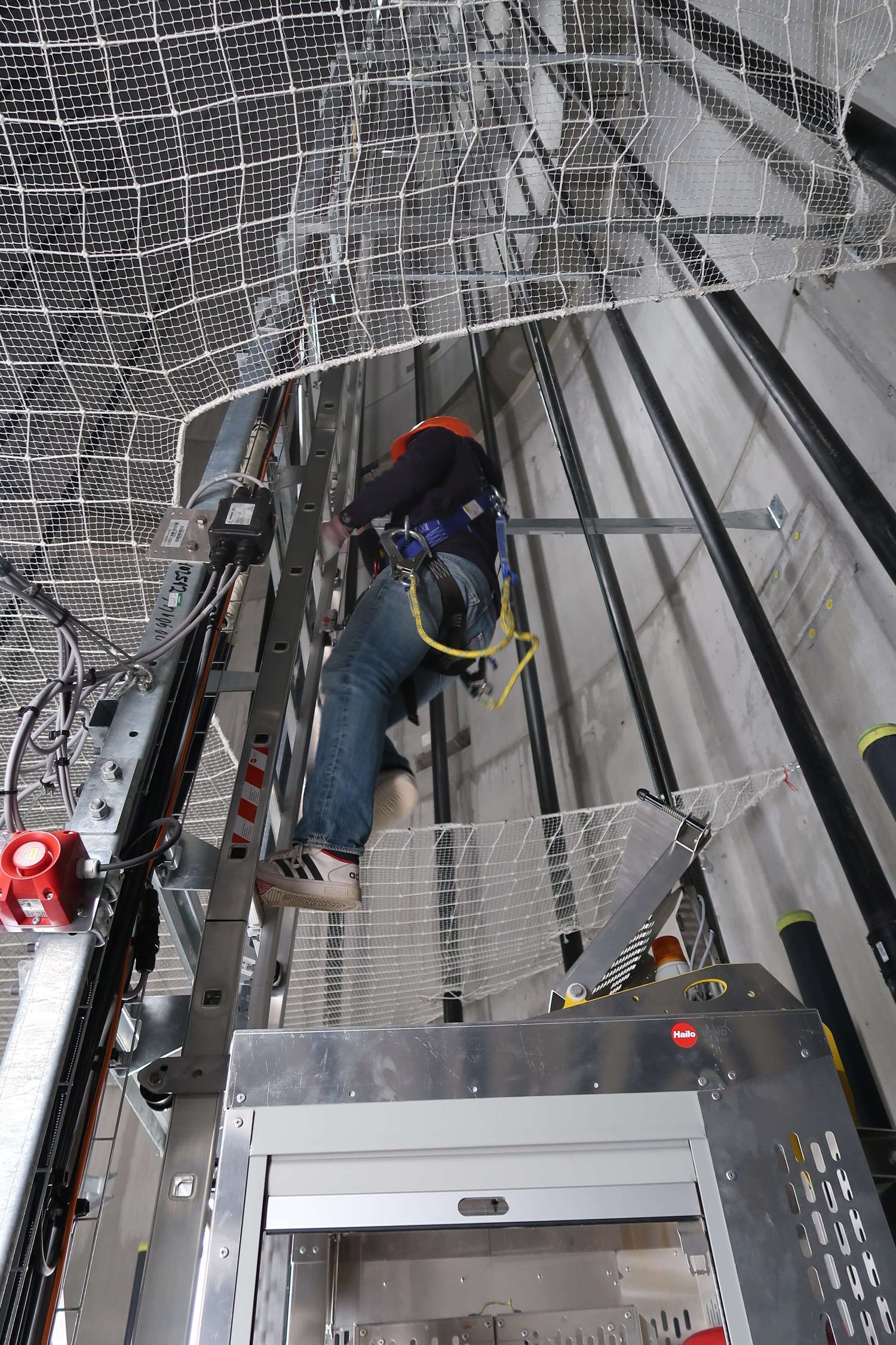 Ein Arbeiter in Sicherheitsausrüstung klettert eine Leiter in einem Aufzugsschacht hinauf, umgeben von Metallstrukturen und Schutznetzen.
