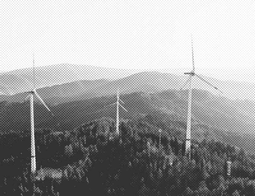 Vier Windräder auf einem bewaldeten Berg mit weiteren Bergen im Hintergrund