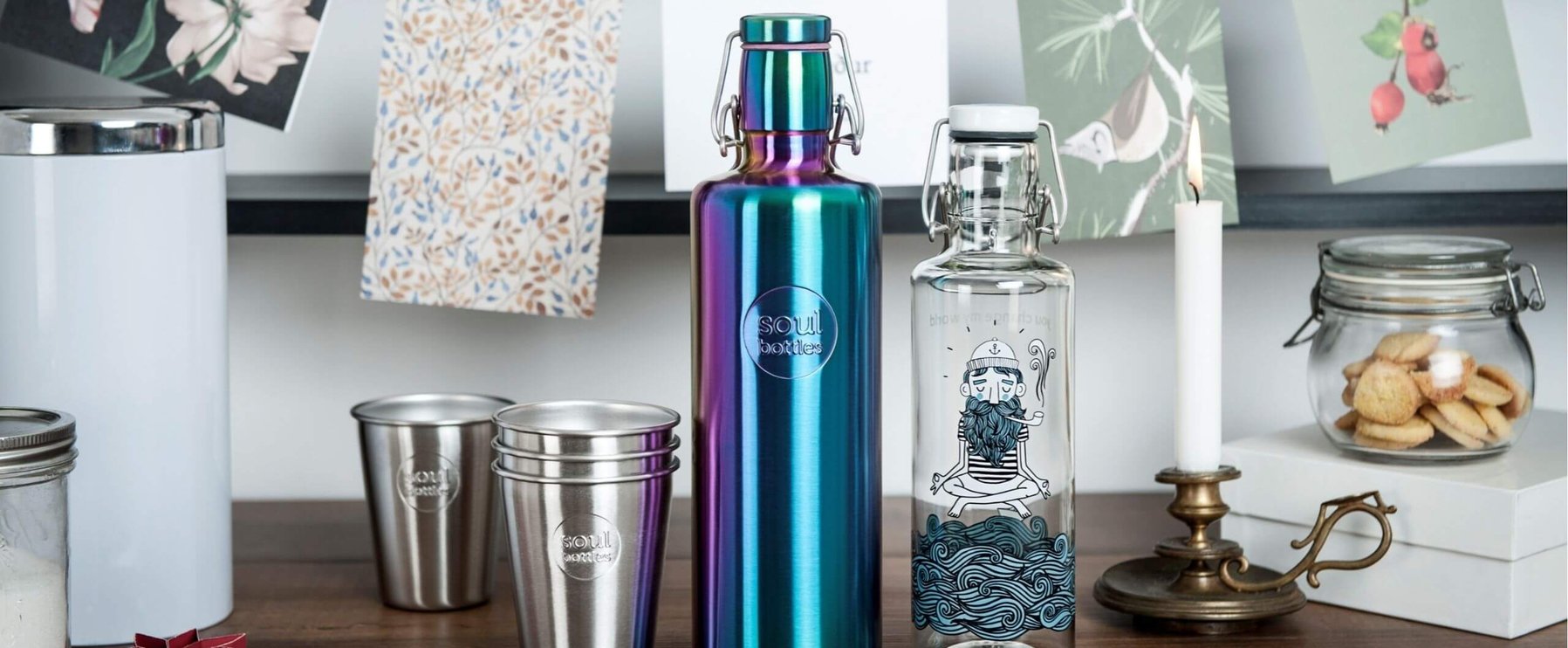 Eine bunte Wasserflasche aus Metall und eine Glasflasche mit einer Illustration stehen auf einem Tisch, umgeben von Metallbechern, einem Kerzenständer, Keksen in einem Glas und hängenden Karten.