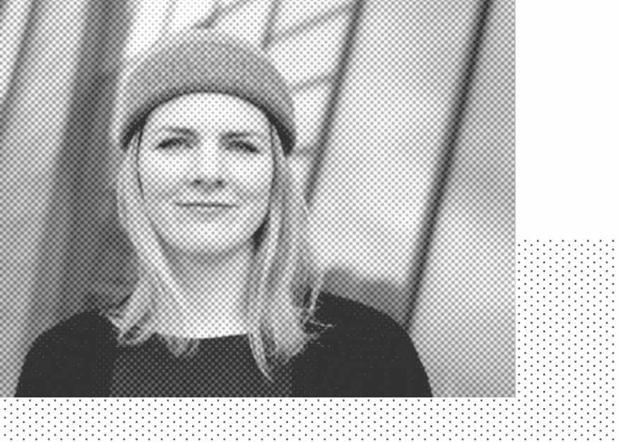 Eine Person mit langen Haaren und einem Beanie auf dem Kopf lächelt leicht und steht vor einem geometrischen Hintergrund. Das Bild ist schwarzweiß.