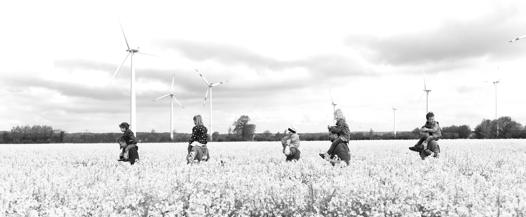 Menschen stehen mit Kindern auf den Schultern in einem Blumenfeld mit Windrädern im Hintergrund.