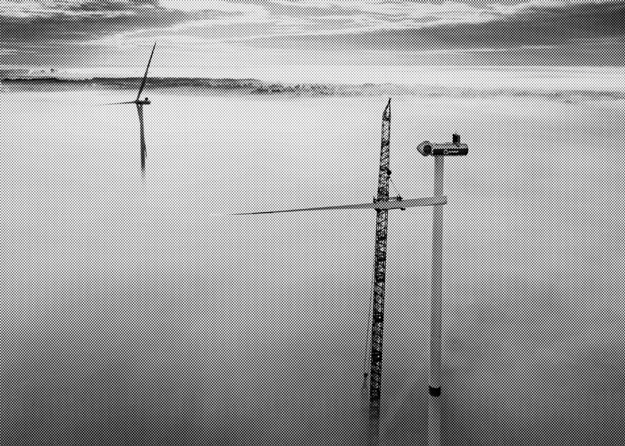 Eine im Bau befindliche Windkraftanlage mit Kran, umgeben von einer nebligen Landschaft. In der Ferne ist eine weitere fertige Windkraftanlage zu sehen.