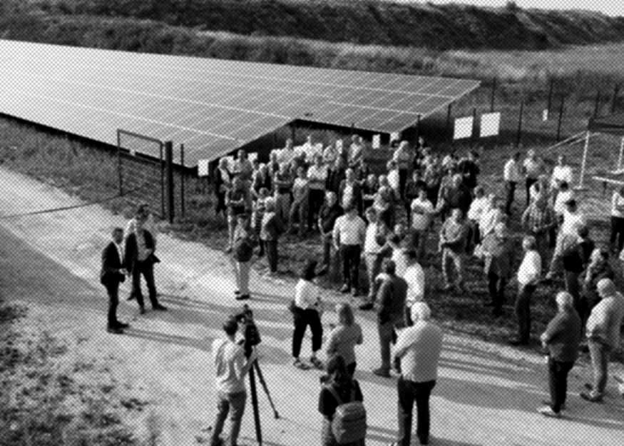 Eine Gruppe von Menschen versammelt sich in der Nähe einer großen Solaranlage. Einige Personen haben Kameras und Ausrüstung dabei, was auf eine Medien- oder offizielle Veranstaltung schließen lässt. Die Szene scheint im Freien an einem sonnigen Tag zu sei
