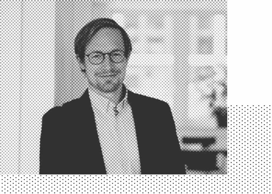 Ein Mann mit Brille und mittellangem Haar, der ein Jackett und ein Hemd trägt, steht im Innenbereich. Das Bild ist in einem Schwarz-Weiß-Halbtonstil gehalten.