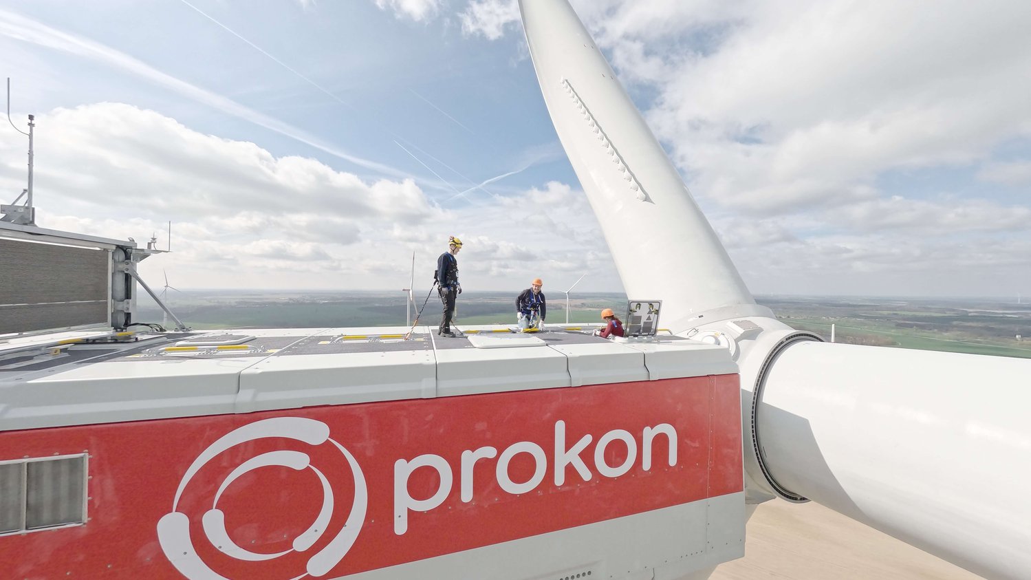 Drei Arbeiter stehen auf einer Windturbine mit der Aufschrift „Prokon“. Vor einem teilweise bewölkten Himmel ist ein Turbinenblatt sichtbar.