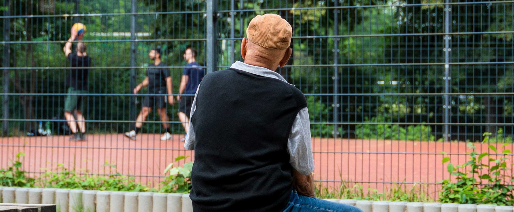 Eine ältere Person mit Mütze beobachtet drei Leute, die auf einem Außenplatz hinter einem Zaun Basketball spielen.