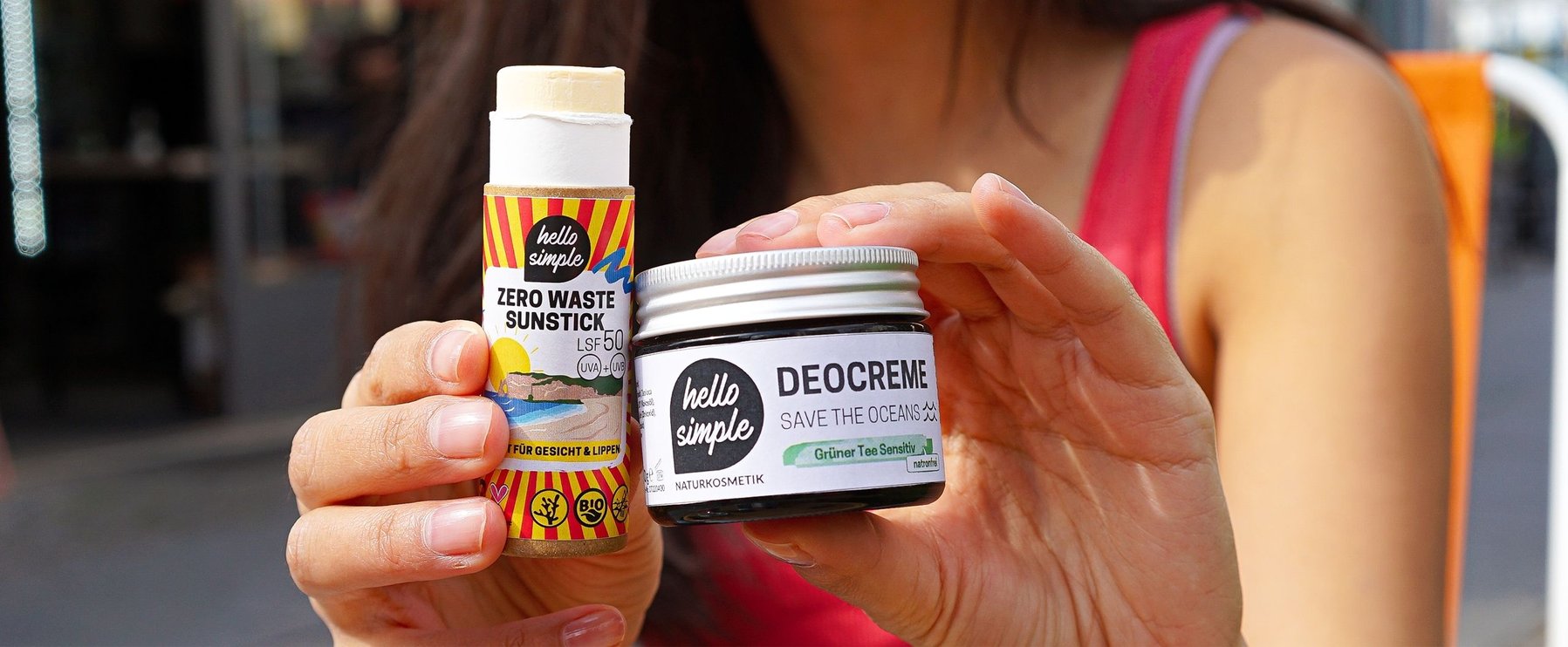 Eine Person hält einen „Zero Waste Sunstick“ und ein Glas „Deocreme – Save the Oceans“ von Hello Simple. Die Produkte sind in umweltfreundlichen Behältern verpackt.