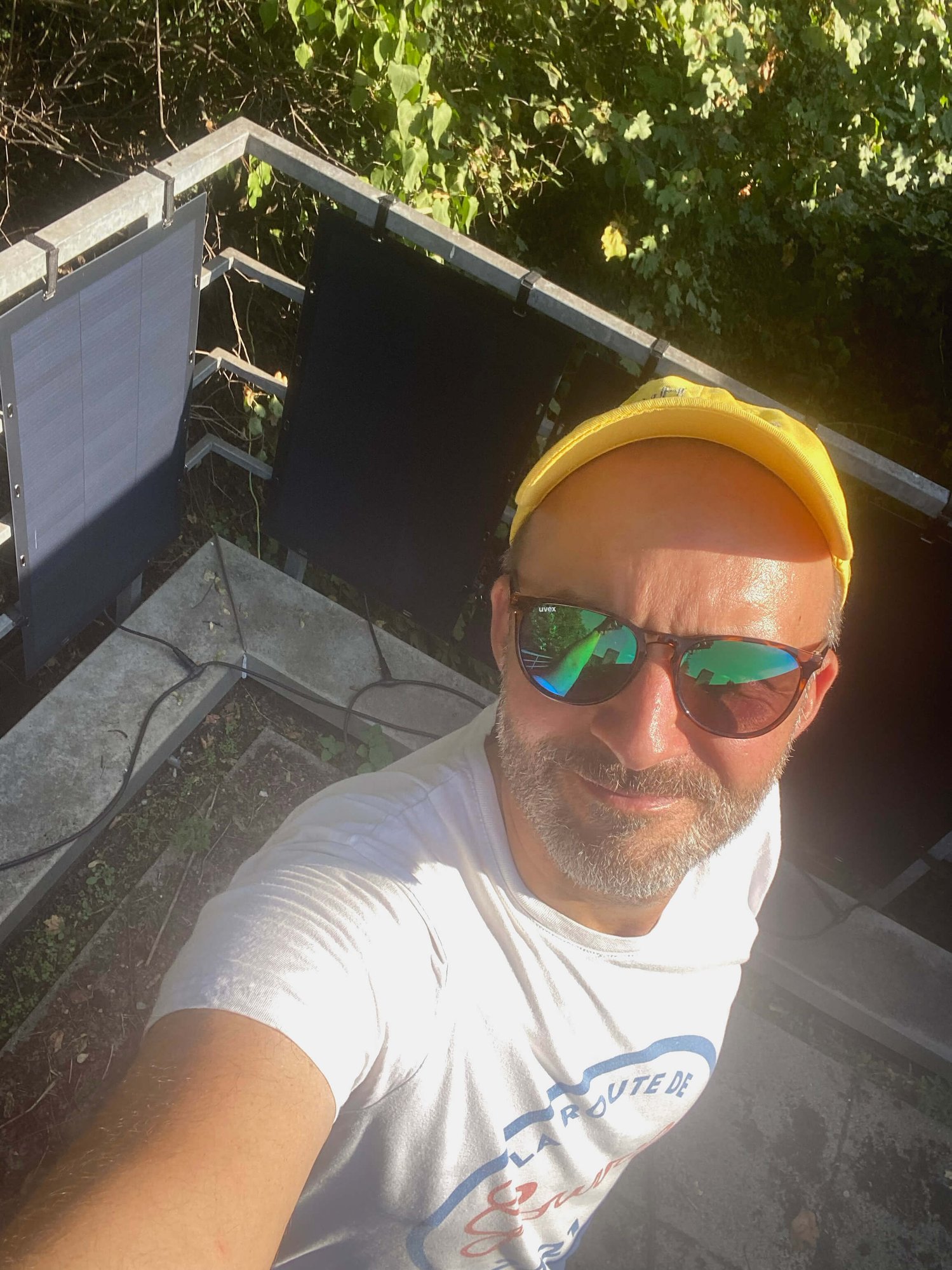 Eine Person mit gelber Mütze und Sonnenbrille macht draußen ein Selfie. Sie steht neben Solarmodulen auf einem kleinen Balkon mit viel Grün im Hintergrund.
