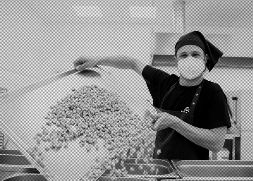 In einer Küche schüttet eine Person mit Maske und Hut Cashewkerne von einem großen Tablett in Behälter.
