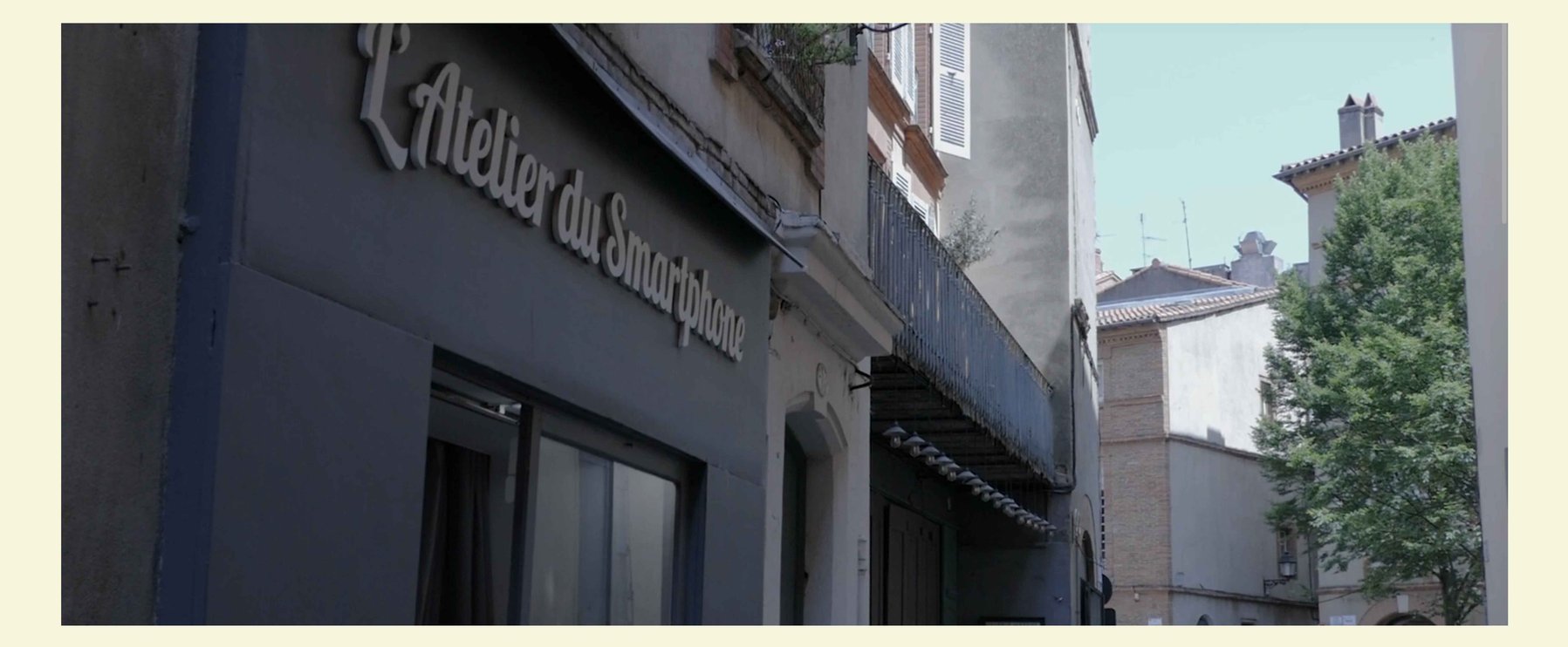 Straßenansicht eines Gebäudes mit einem Schild mit der Aufschrift „L'Atelier du Smartphones“. Im Hintergrund sind Bäume und Dächer unter einem klaren Himmel zu sehen.