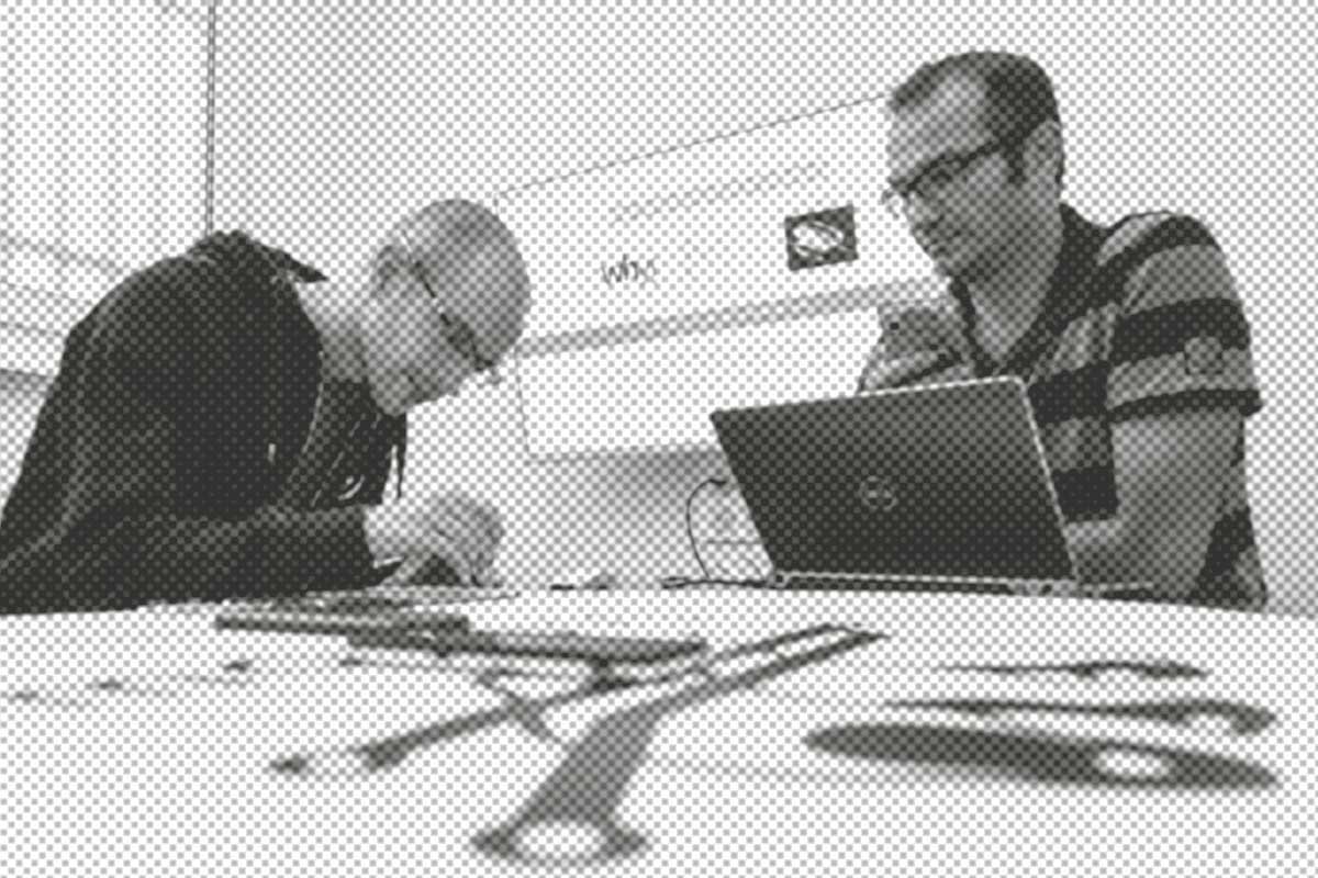 Zwei Personen arbeiten an einem Tisch mit Skizzen und Werkzeug. Eine Person nutzt einen Laptop, die andere konzentriert sich auf eine Aufgabe.