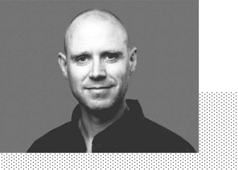 Schwarz-Weiß-Porträt einer Person mit rasiertem Kopf, die ein dunkles Hemd mit Kragen trägt und vor einem grauen Hintergrund leicht lächelt.