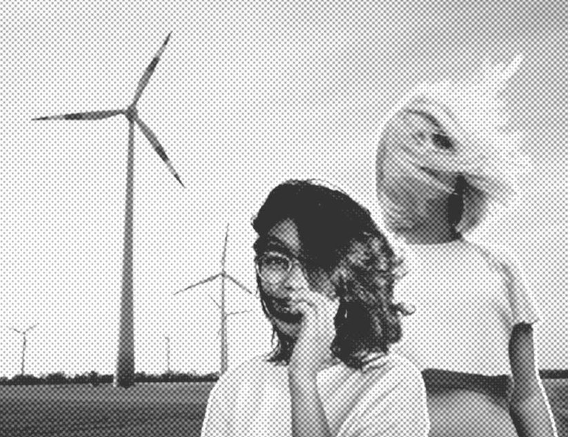 Zwei Personen stehen auf einem Feld mit Windrädern im Hintergrund. Eine Person scheint sich ins Gesicht zu fassen, während die andere in den Himmel blickt.