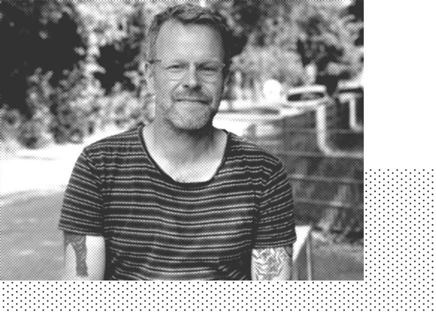 Mann mit Brille und Tattoos, trägt ein gestreiftes Hemd, sitzt im Freien mit Bäumen im Hintergrund.