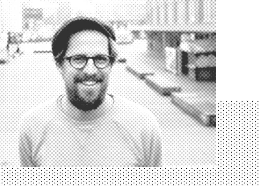 Ein Mann mit Brille und Hut steht lächelnd auf der Straße. Das Bild hat einen Halbtoneffekt.