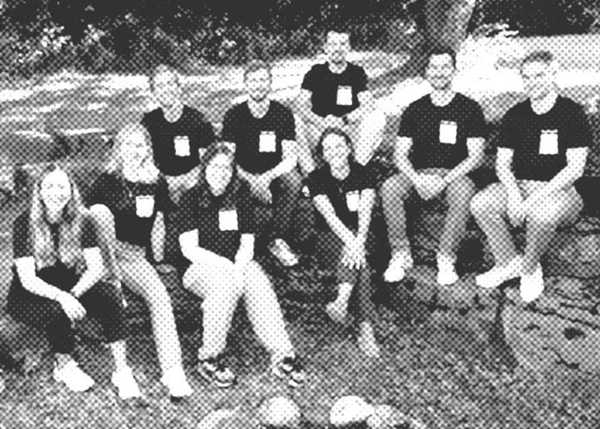 Ein Schwarzweißfoto einer Gruppe von zehn Personen, die im Gras sitzen und knien. Alle tragen passende dunkle T-Shirts mit sichtbaren Namensschildern. Im Hintergrund sind Bäume und ein Schatten zu sehen.