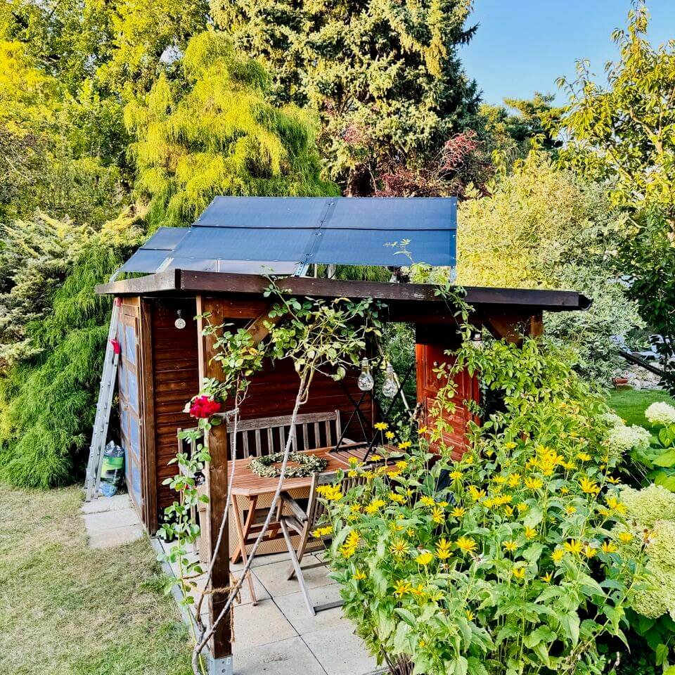 Ein kleines Gartenhäuschen aus Holz mit einem Dach aus Solarmodulen ist von üppigem Grün und gelben Blumen umgeben. An der Vorderseite stehen unter einer Pergola eine Bank und ein Stuhl.