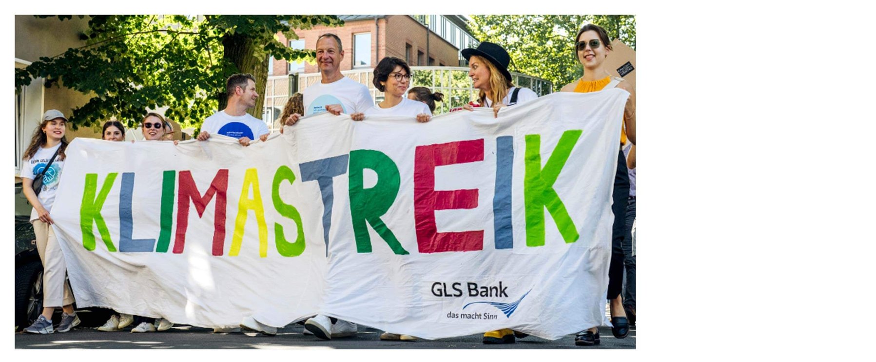 Menschen halten während einer Klimastreik-Veranstaltung im Freien vor Bäumen und einem Gebäude ein Transparent mit der Aufschrift „Klimastreik“ in bunten Buchstaben.