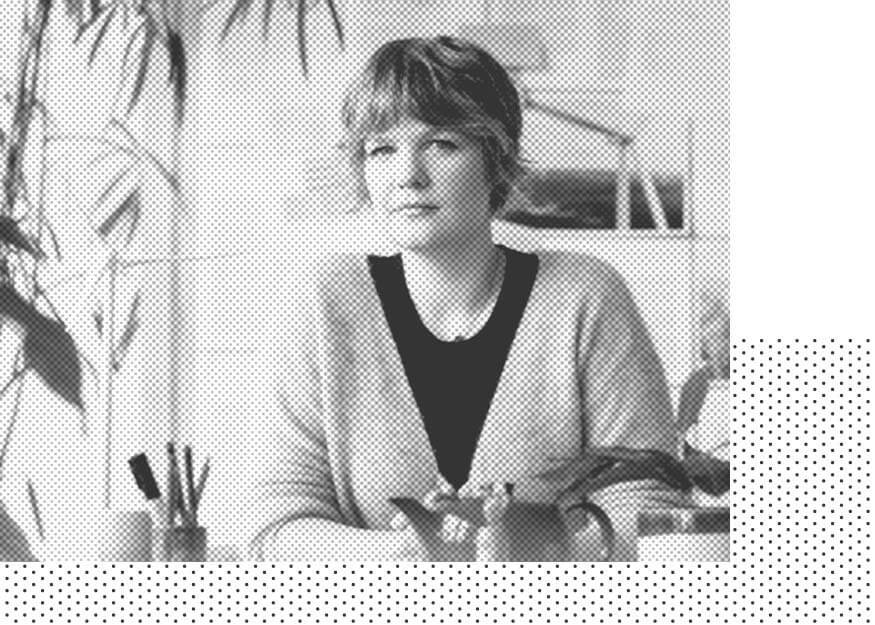 Eine Frau mit kurzen Haaren sitzt an einem Schreibtisch, umgeben von Pflanzen und Schreibwaren. Das Bild ist schwarz-weiß.