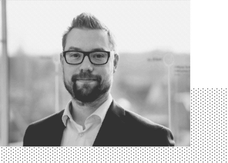 Schwarzweißfoto eines Mannes mit Brille, Bart und kurzen Haaren, der ein Jackett und ein Hemd trägt und lächelt. Der Hintergrund ist unscharf.