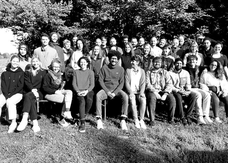 Ein Schwarzweiß-Gruppenfoto von 38 Personen, die draußen auf dem Gras sitzen und stehen, mit Bäumen im Hintergrund.