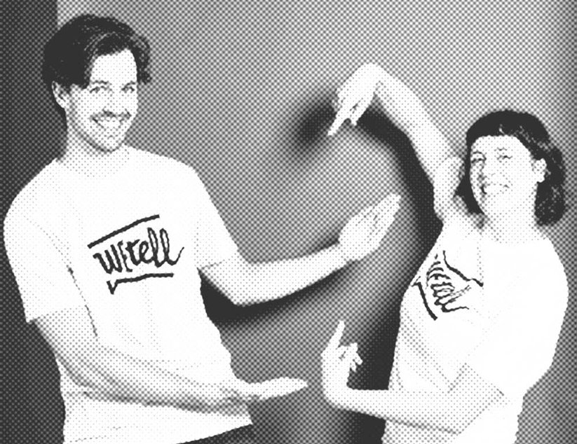 Zwei Personen in passenden WEtell-T-Shirts posieren begeistert und zeigen auf den Text auf ihren Shirts vor einem schlichten Hintergrund.