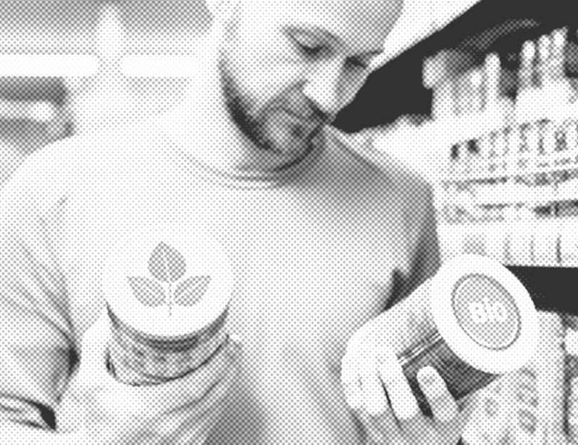 Ein Mann steht im Supermarkt und betrachtet zwei Dosen, die er in den Händen hält. Auf der einen steht "Bio", auf der anderen ist ein Label mit einer stilisierten Pflanze zu sehen.