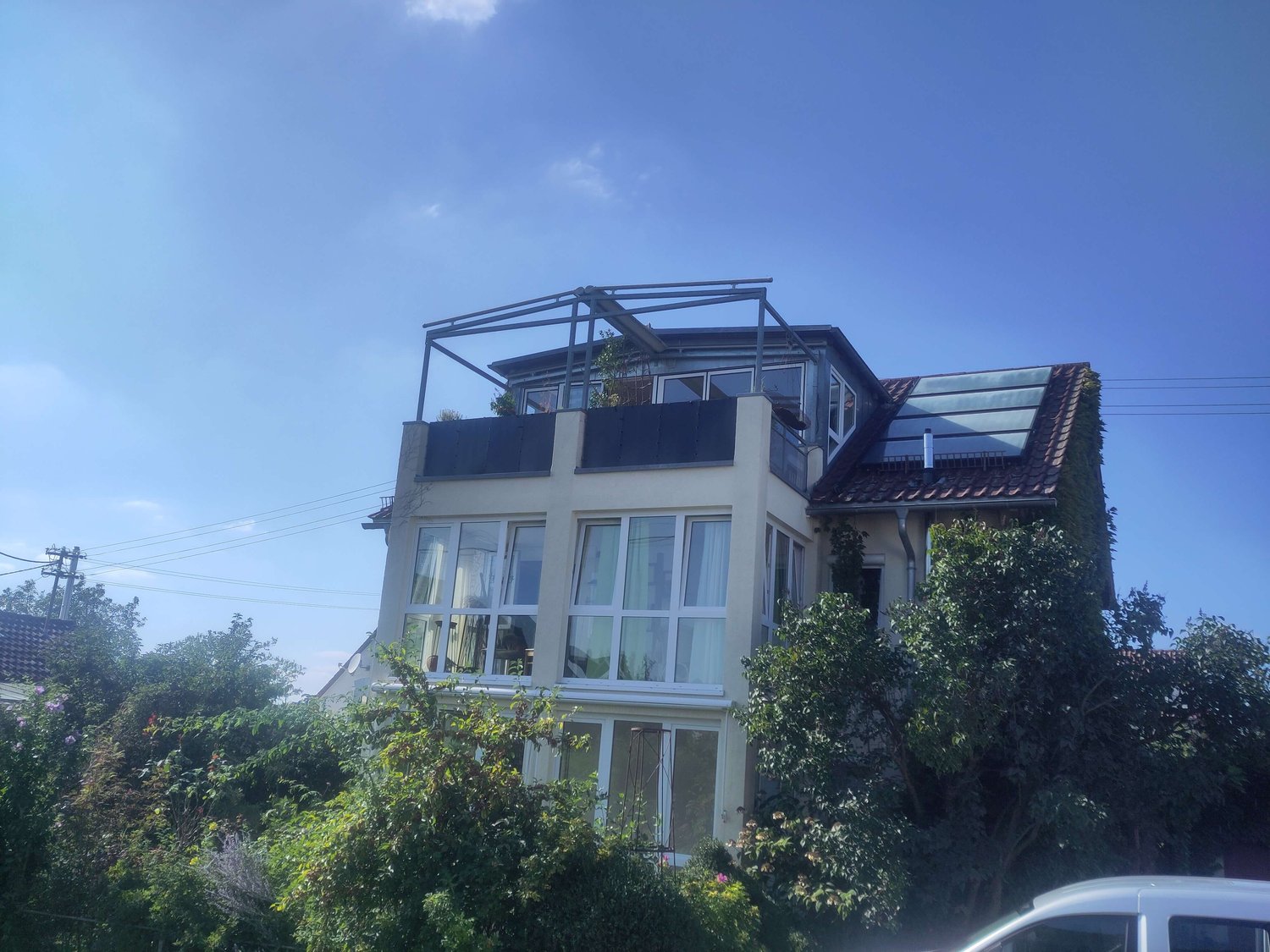 Ein mehrstöckiges Haus mit großen Fenstern, einer Pergola auf dem Dach und umgebendem Grün unter einem klaren blauen Himmel.