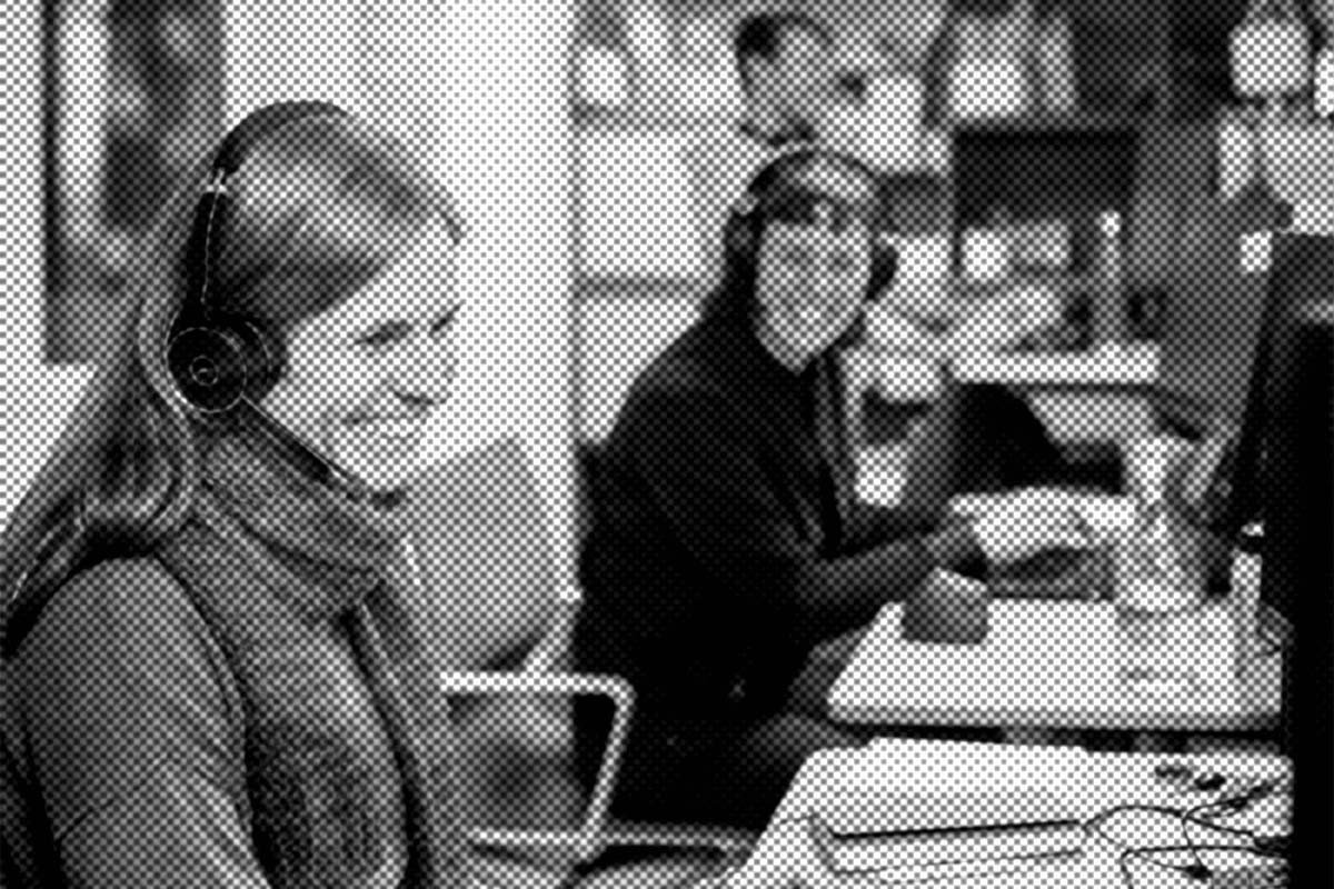 Zwei Personen mit Headsets sitzen an Schreibtischen in einem Büro und lächeln. Im Hintergrund sind Büroartikel zu sehen, und eine dritte Person arbeitet ebenfalls am Computer.