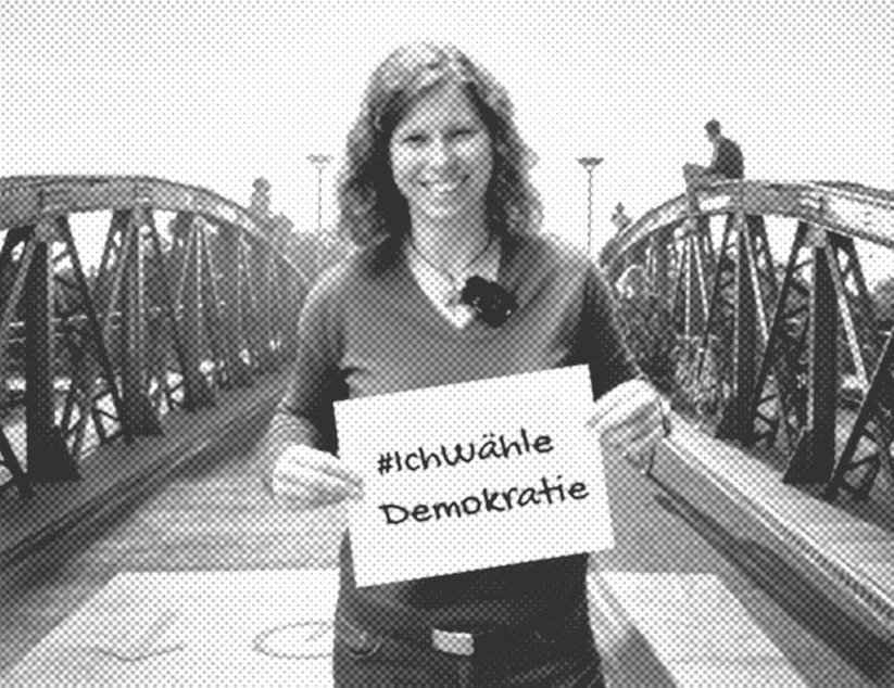 Eine lachende Frau steht auf einer Fahrradbrücke und hält ein Schild vor sich auf dem steht "Ich wähle Demokratie".