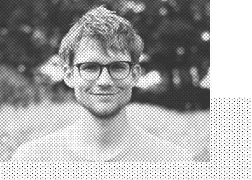Eine junge Person mit kurzen Haaren und Brille lächelt im Freien in die Kamera. Das Bild ist schwarz-weiß mit einem Halbtonmuster.