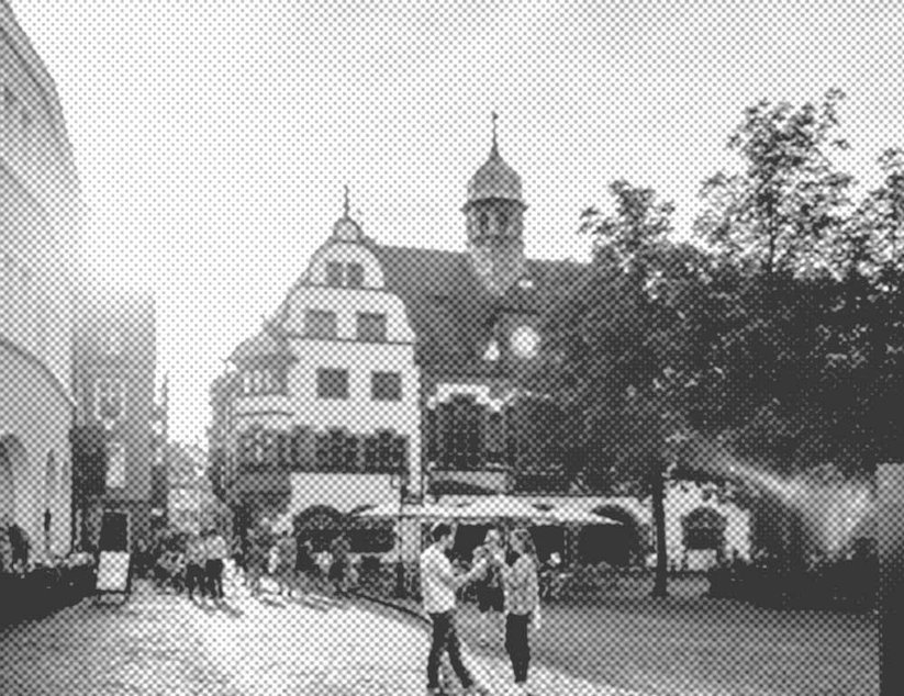 Ein historischer europäischer Stadtplatz mit Passanten. Ein Fachwerkhaus mit Uhrturm und Café-Sitzplätzen ist sichtbar. Bäume säumen die Kopfsteinpflasterstraße.