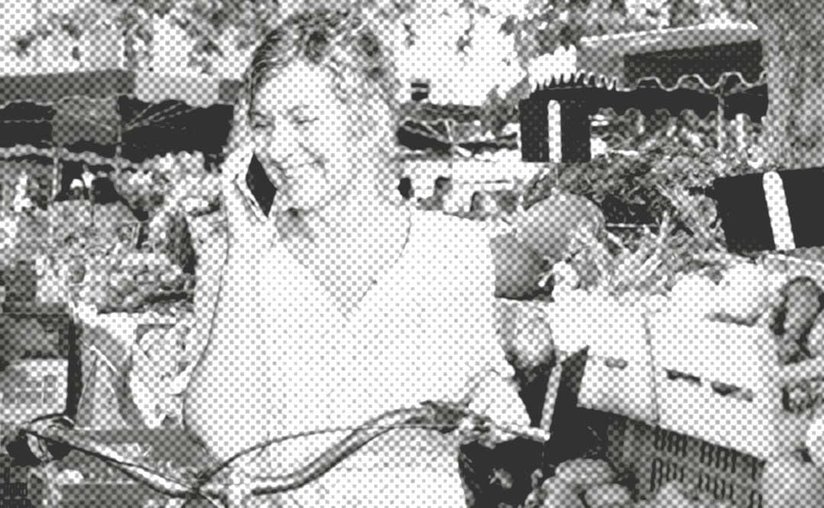 Eine Frau steht lächelnd auf einem Markt im Freien und telefoniert, während um sie herum frische Produkte zu sehen sind.