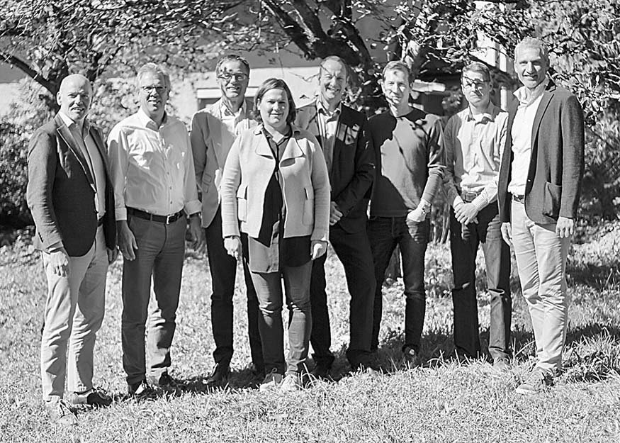 Gruppe von neun Personen, die im Freien auf Gras stehen, mit Bäumen und einem Gebäude im Hintergrund, auf einem Schwarzweißfoto.