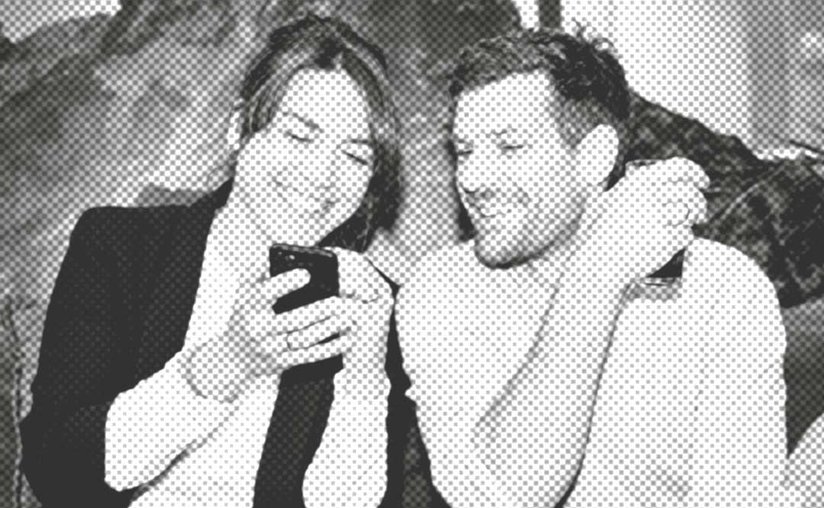 Zwei Menschen sitzen zusammen auf einer Couch, beide schauen auf ein Smartphone und lächeln.