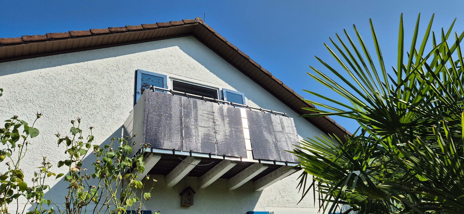 Ein Haus mit einem kleinen, mit Solarmodulen behangenen Balkon, blauen Fensterläden und einem schrägen Dach. Im Vordergrund sind Palmblätter und Pflanzen unter einem klaren blauen Himmel zu sehen.