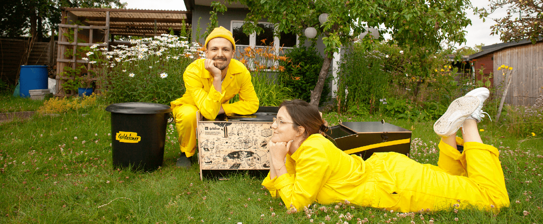 Zwei Personen in passenden gelben Outfits posieren auf einer Rasenfläche mit einer schwarzen Tonne und einer dekorierten Kiste, umgeben von Grün und Blumen, vor einer kleinen Gartenlaube.