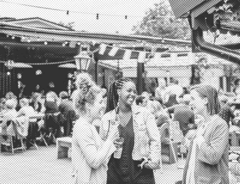 Drei junge Frauen stehen im Vordergrund, reden miteinander und lachen. Im Hintergrund sieht man ein Fest in einem großen Hof, dekoriert mit Wimpeln, unter denen Menschen an großen Tischen.