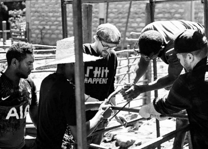Fünf Personen arbeiten gemeinsam an einer Metallkonstruktion im Freien in einem sonnenbeschienenen Bereich. Eine Person trägt einen Hut, eine andere eine Brille und mehrere halten ein Stück Metall.