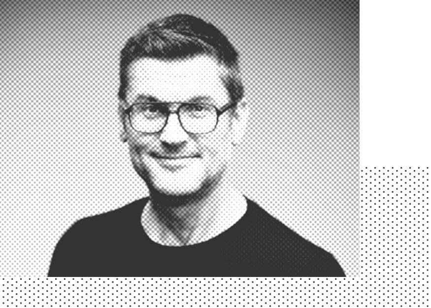 Schwarz-weißes Halbtonporträt eines Mannes mit Brille und kurzen Haaren, der ein dunkles Hemd trägt und in die Kamera lächelt.