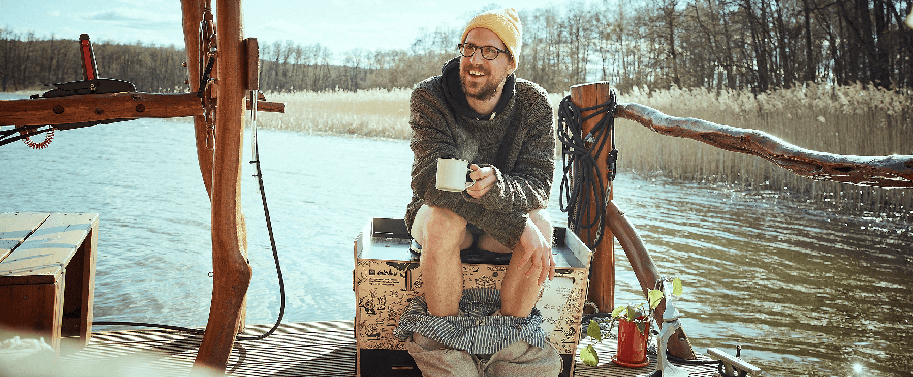 Eine Person sitzt mit heruntergelassenen Hosen auf deinem Trockenklo auf einem Floß im Wasser, hält einen Becher in der Hand.