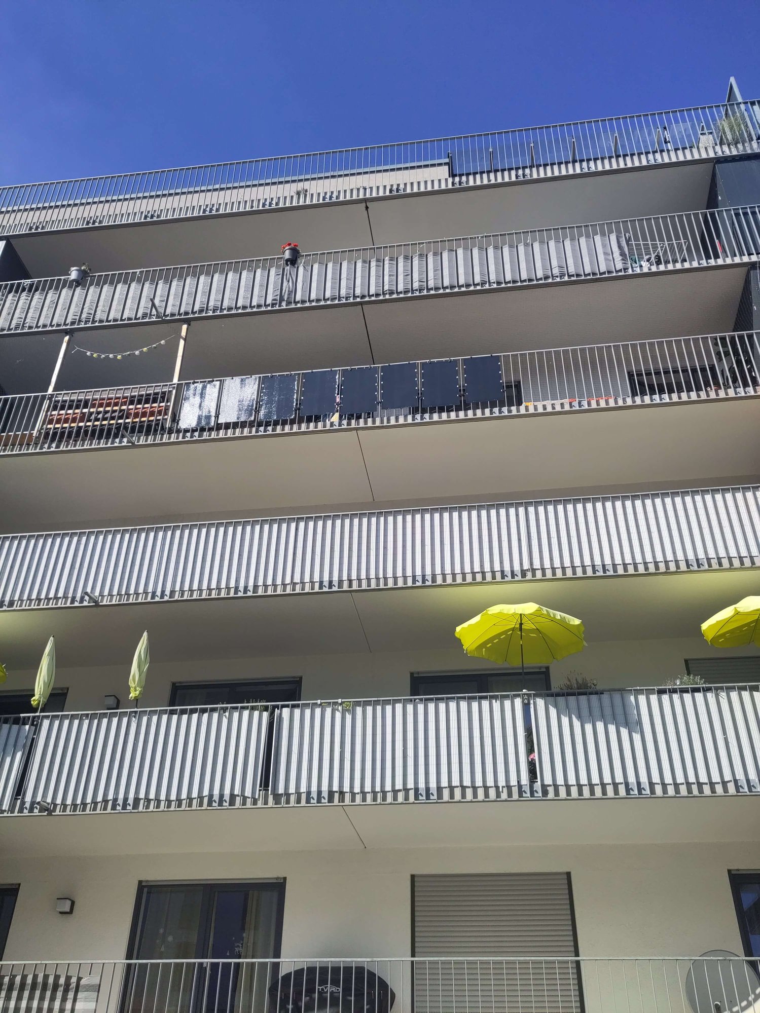 Mehrstöckiges Gebäude mit mehreren Balkonen, zwei davon mit gelben Sonnenschirmen und einer mir Solarpanelen behangen. Der Himmel ist klar und blau.