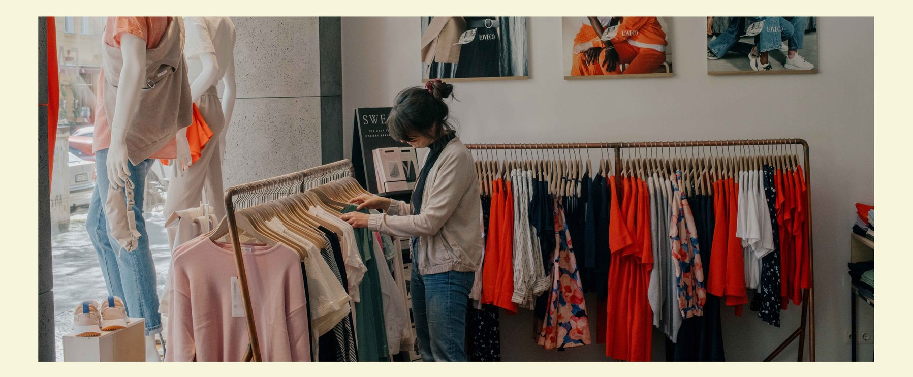 Eine Frau stöbert in einem Geschäft durch Kleiderständer mit Fair-Fashion-Kleidung. Modeposter schmücken die Wände, während natürliches Licht durch ein Fenster hereinströmt.
