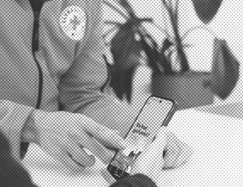 Zwei Personen sitzen am Tisch, eine ist nur von hinten zu sehen und hält ein Handy in der Hand, die andere trägt eine Jacke der Bahnhofsmission und zeigt auf das Handy.